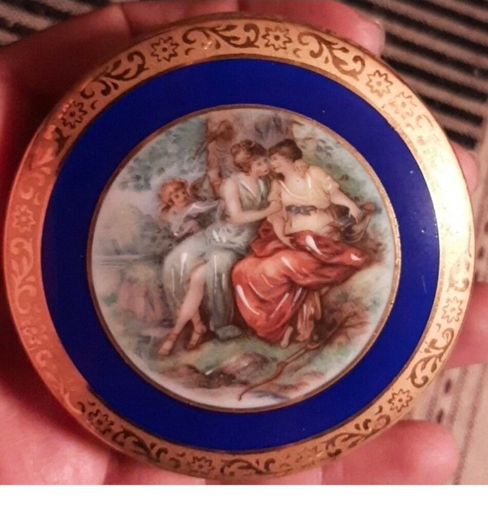 Round compact lid with a classical scene of two women and a cherub, framed by a cobalt band and gold scroll border